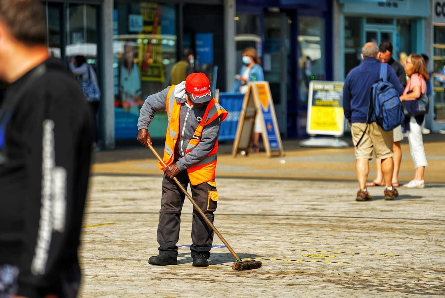 A colourful image showing a high street focusing on one man in a hi vis jacket sweeping the bricked floor on a dry cold day.